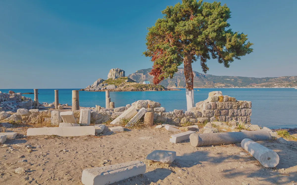 Kos adasında deniz kenarında antik kalıntılar, uzakta küçük bir ada ve şapel manzarası.