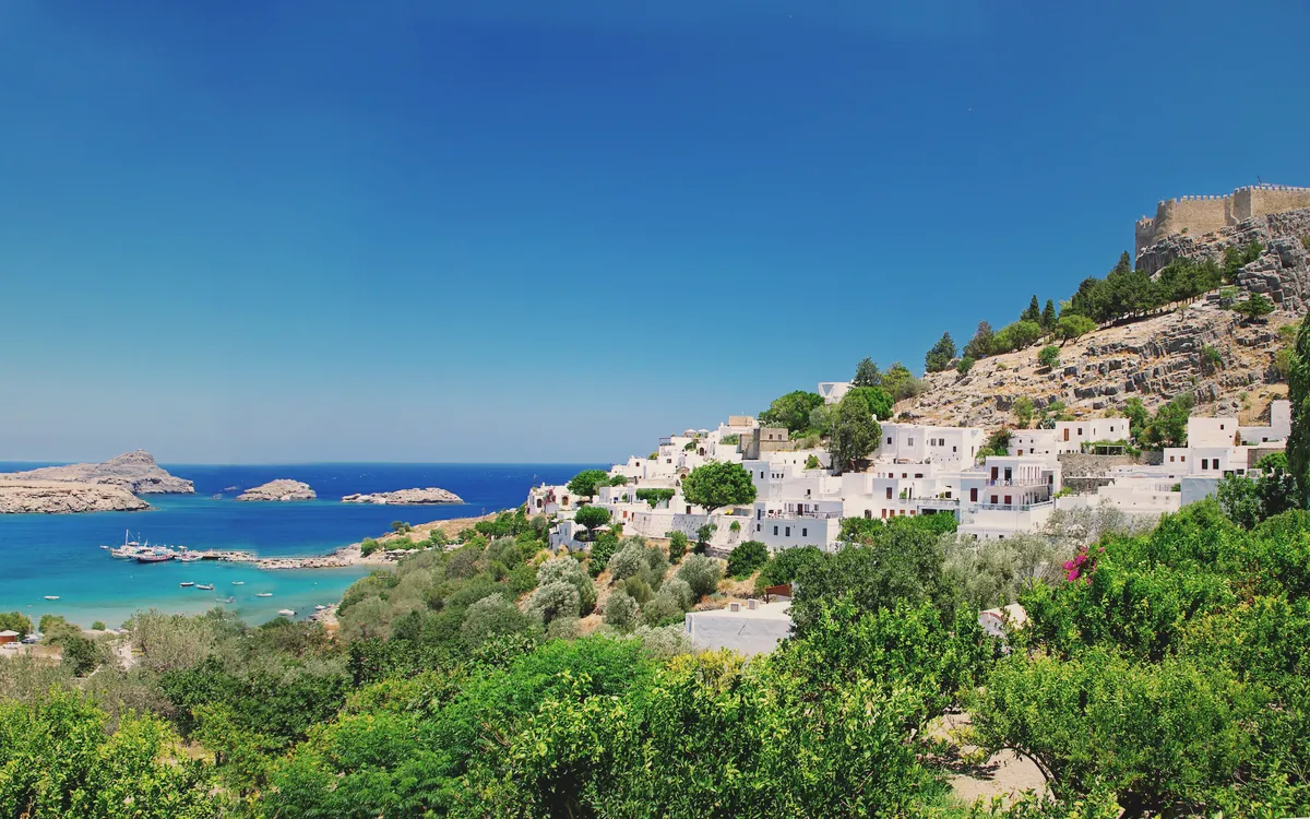 Rodos'taki Lindos köyünün beyaz evleri, masmavi denizi ve tepedeki antik akropolisi gösteren panoramik bir manzara.