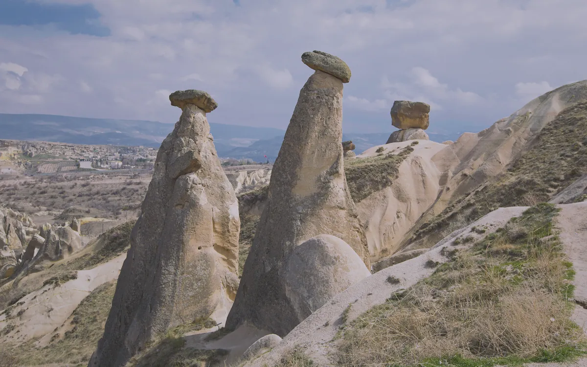 Kapadokya'da, tepeleri şapkalı üç büyük peri bacası ve uzakta bir kasaba görünen vadi manzarası.